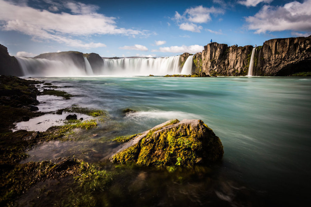 Panoramisch uitzicht op de Godafoss-waterval in IJsland met turquoise water, rotsen en een blauwe lucht.
