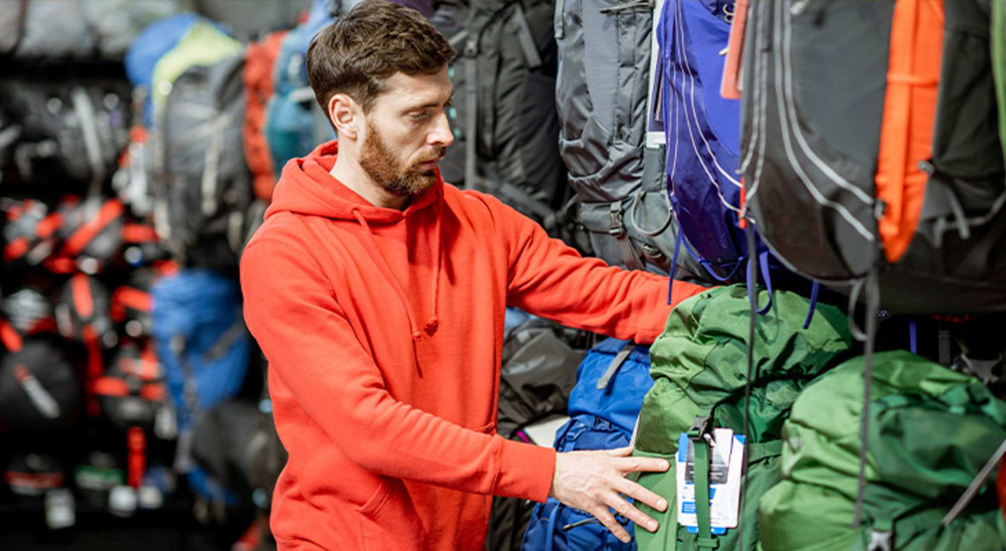 Backpack kopen Man in rode hoodie kiest een trekkingrugzak in een outdoorwinkel.