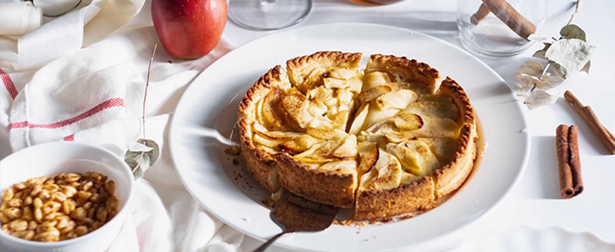 Appeltaart met kaneel op een wit bord, met appel en ingrediënten op de achtergrond.