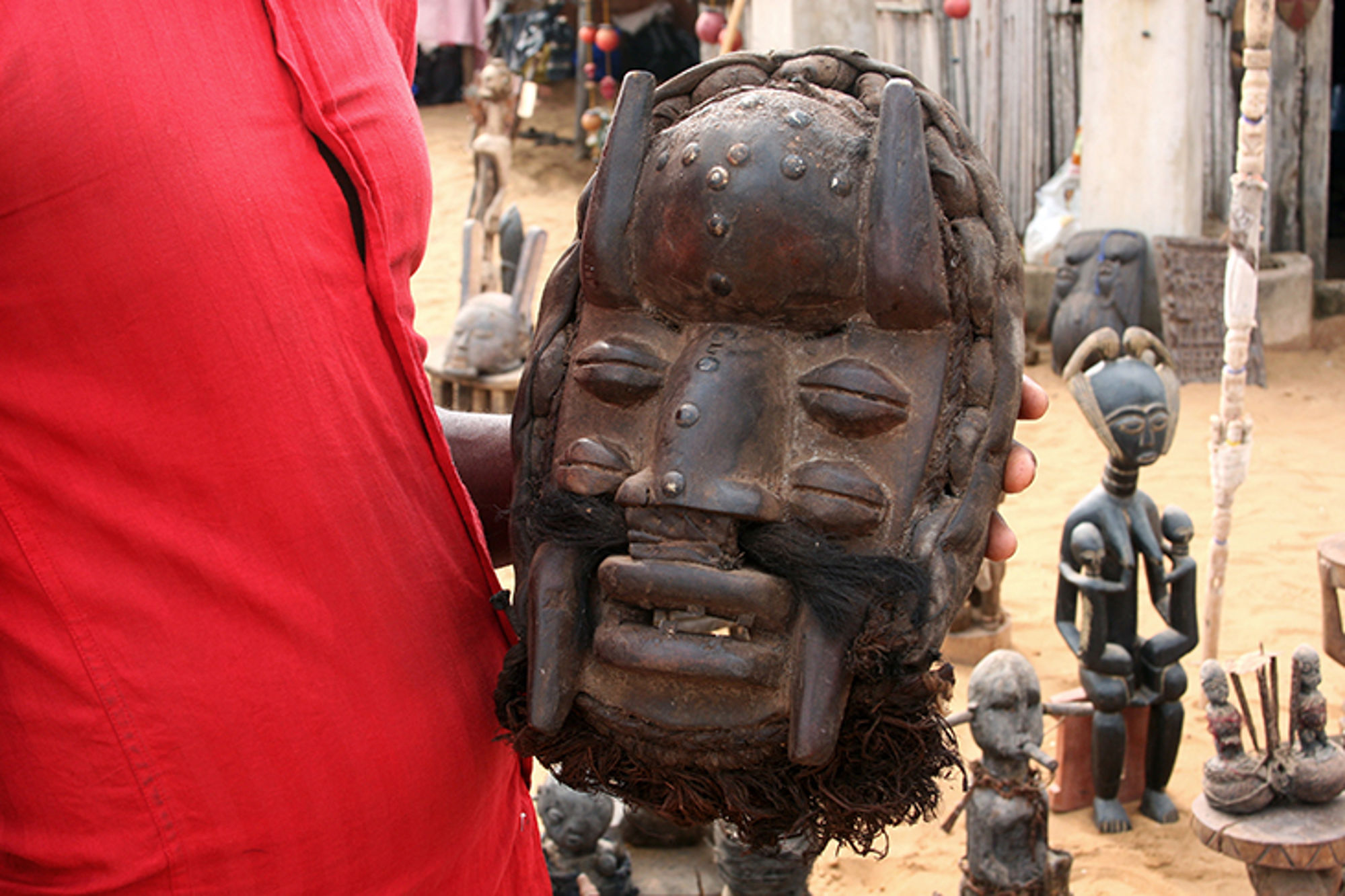 afrikaanse kunst - houten beeld in benin Houten ceremonieel masker met uitgesneden gezicht, vastgehouden op een markt met traditionele beeldjes op de achtergrond.