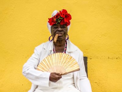 Cubaanse vrouw in witte kleding met rode bloemen in haar hoofddoek, sigaar en waaier tegen gele muur.