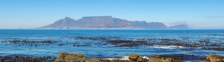 Uitzicht op de Tafelberg en Kaapstad vanaf Robbeneiland, met rotsachtige kust en zee op de voorgrond.