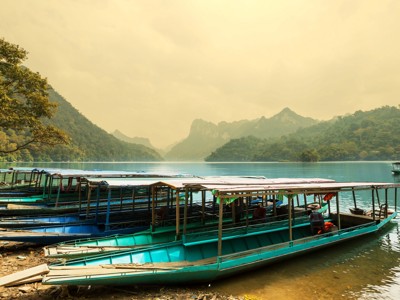Longtailboten aan de oever van het Ba Be-meer in Vietnam, met groene bergen op de achtergrond.