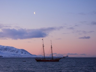 Zeilschip Noorderlicht voor de kust van Spitsbergen bij schemering, met besneeuwde bergen en een sikkelmaan.