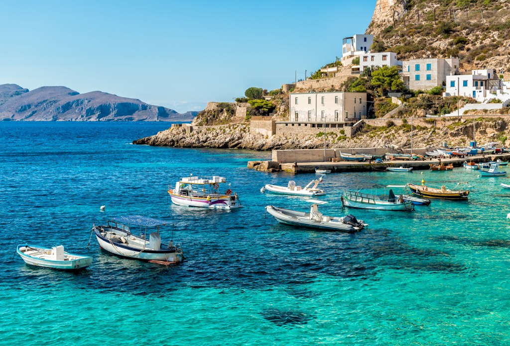 Kustdorp aan de baai in West-Sicilië met vissersbootjes in helderblauw water en huizen tegen de rotswand.
