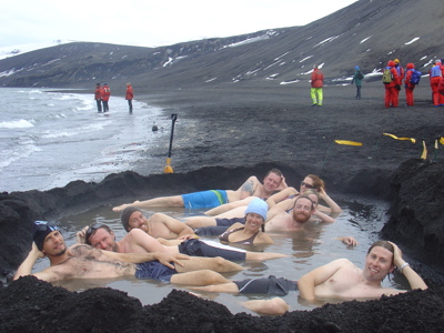 Reizigers ontspannen in een warmwaterpoel op een zwart zandstrand in Antarctica, met besneeuwde heuvels op de achtergrond.