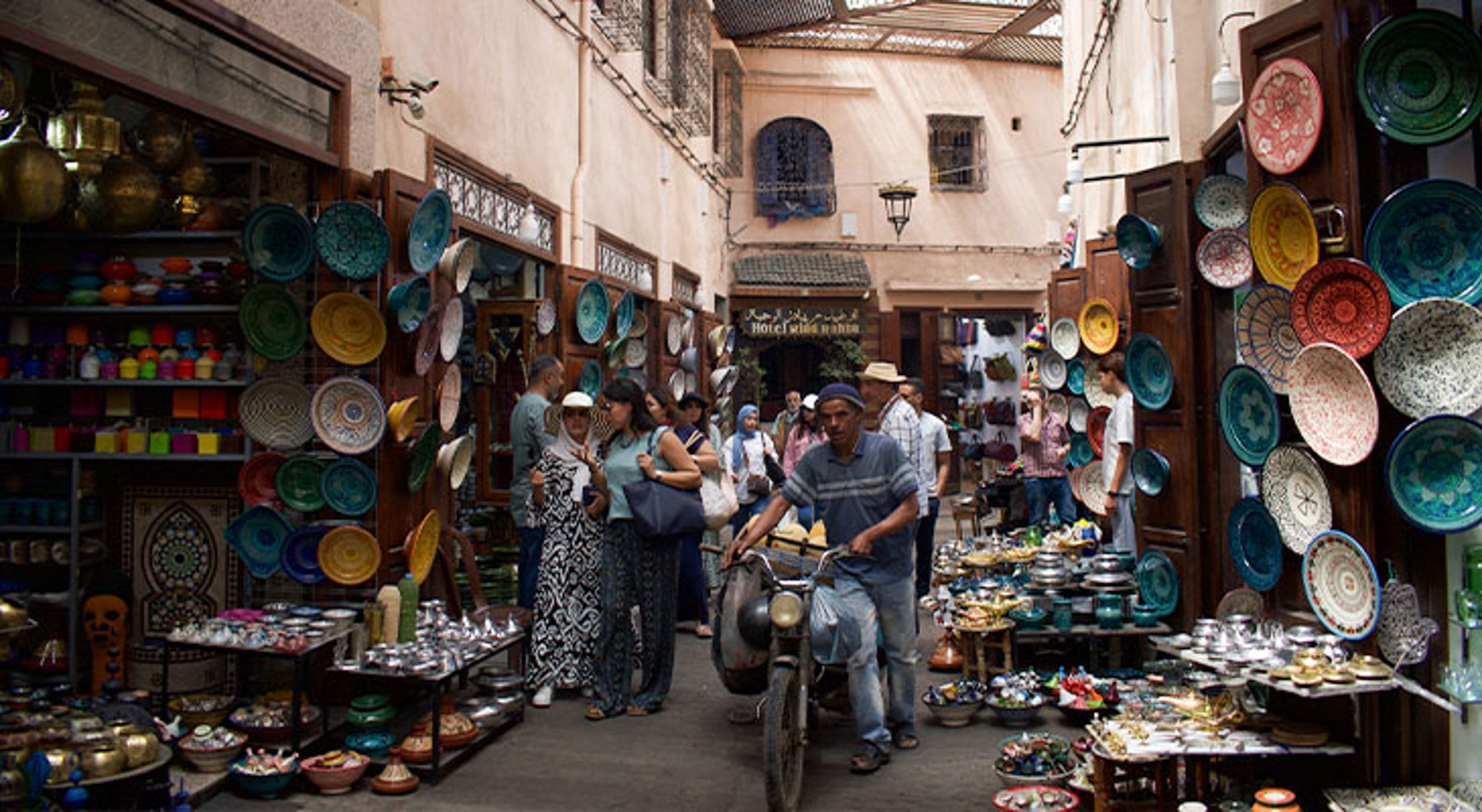 Marrakech Drukke souk in Marokko met kleurrijk aardewerk en schalen langs smalle steeg, terwijl mensen en een fietser passeren.