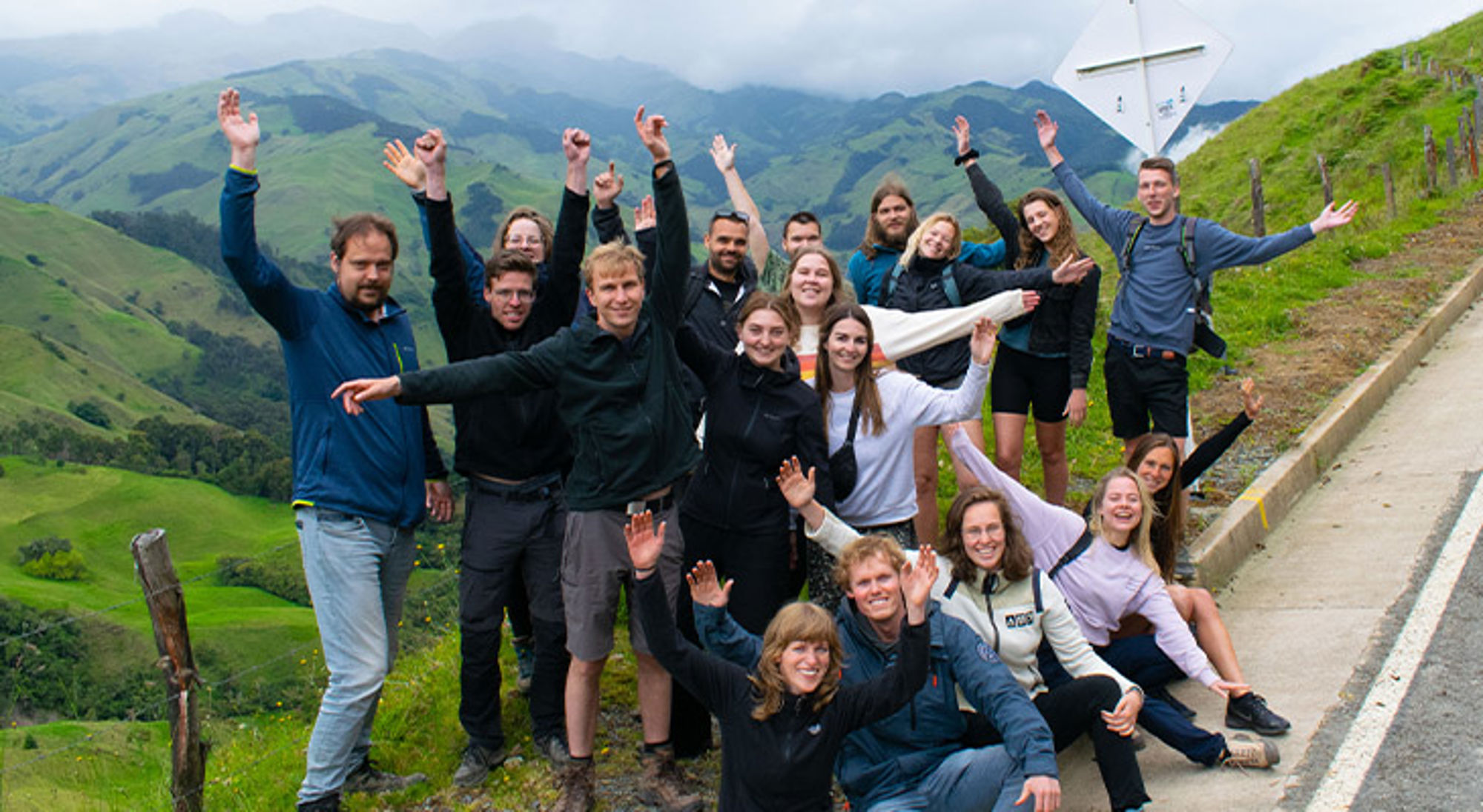 Groep reizigers poseert langs een bergweg met groene heuvels en wolken in de achtergrond.