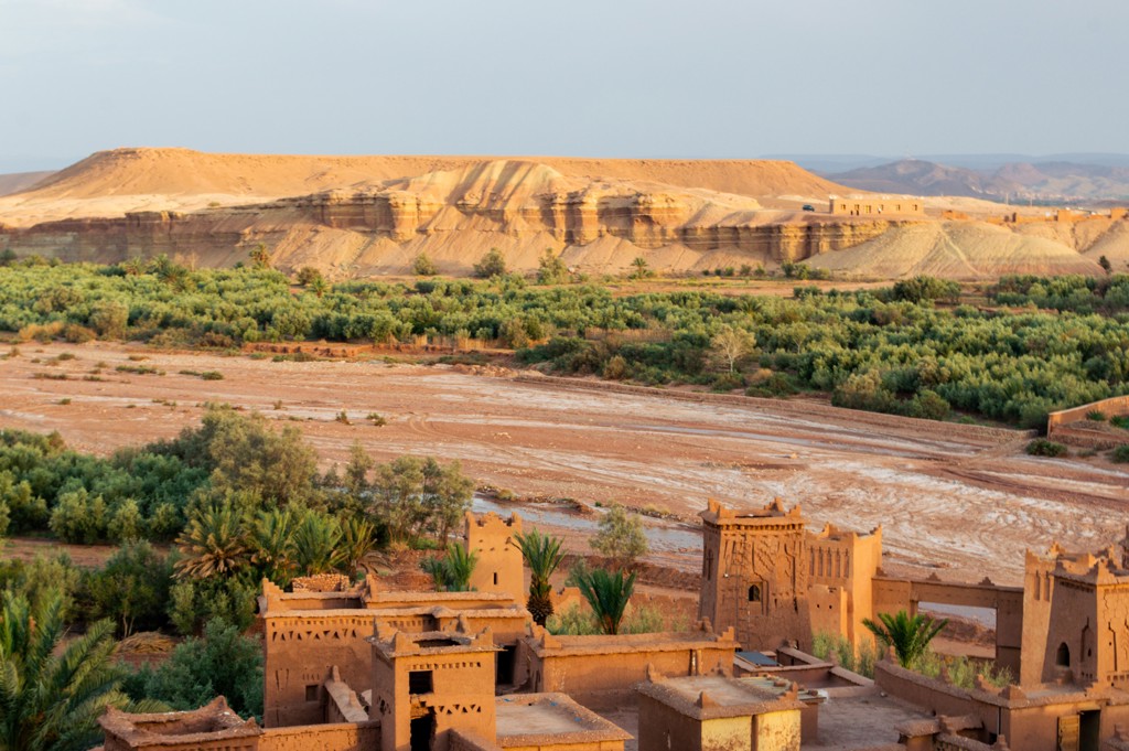 Leemkleurige kasbah aan een groene oase met rivierbedding en rotsplateau, tijdens Familiereis Marokko Hoogtepunten.