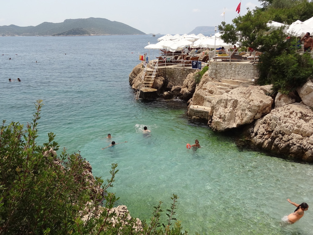 Zwemmers in een heldere baai aan de Turkse Turquoise Kust, met rotsen en strandterras met parasols.