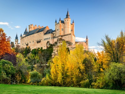 Alcázar van Segovia, kasteel op een rots boven herfstkleurige bomen en een groene weide.
