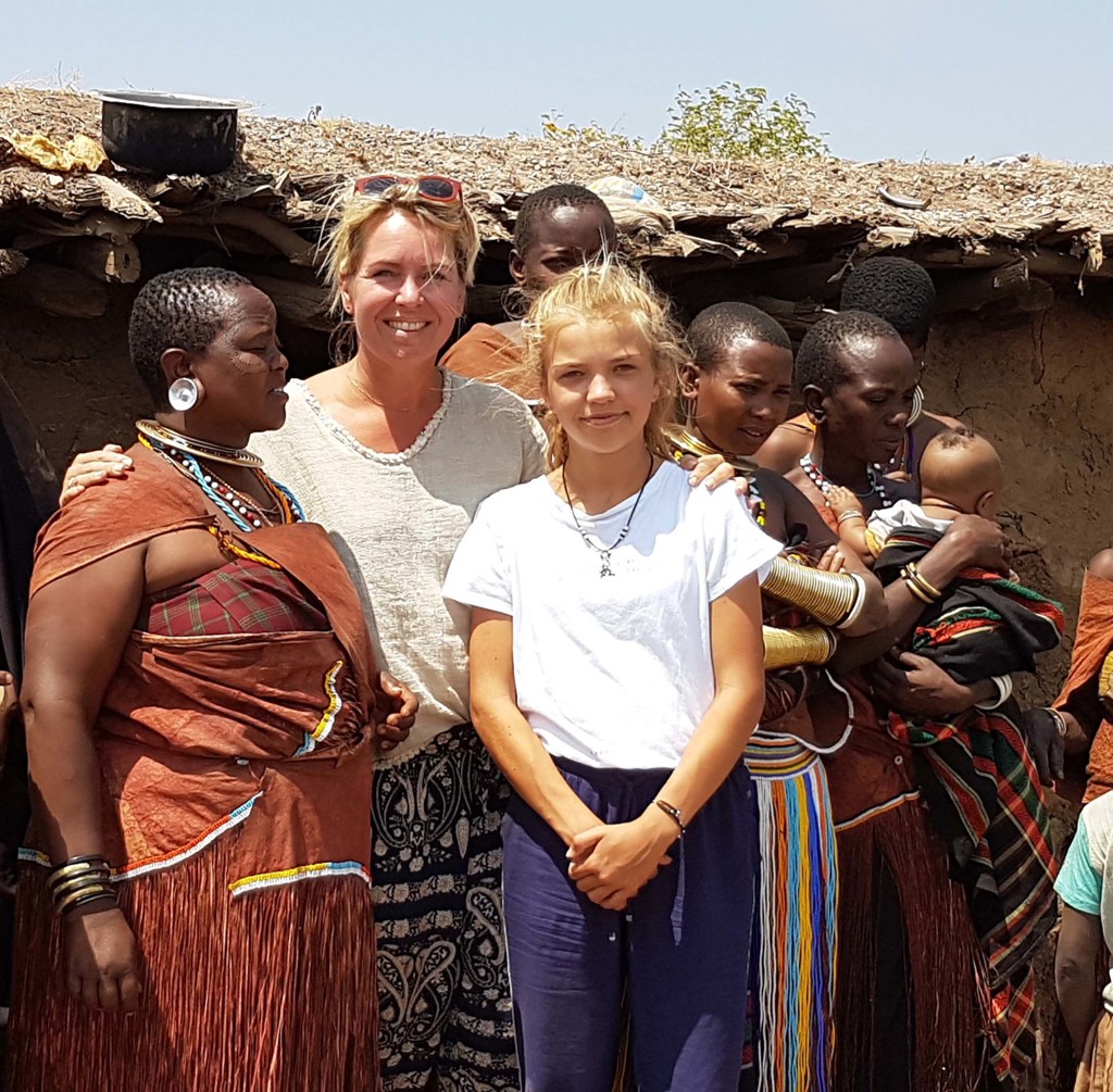 Gezin op familiereis in Tanzania poseert met lokale vrouwen in traditionele kleding bij een hut.