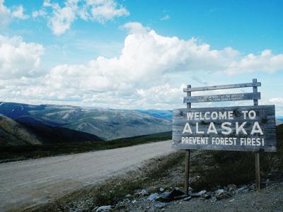 Welkom-bord bij de grens van Alaska langs een onverharde weg, met bergen op de achtergrond.