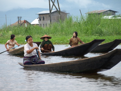 Vrouwen peddelen in houten boten over Inle Lake in Myanmar, langs riet en paalwoningen.