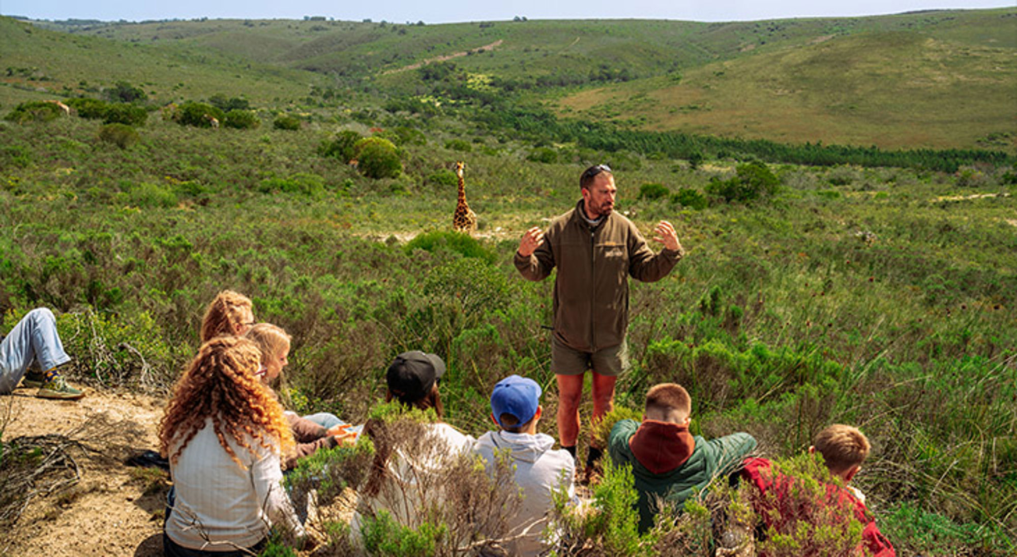 Uitleg ranger Zuid-Afrika Gids vertelt kinderen over safari in een groen natuurgebied, met een giraffe op de achtergrond.