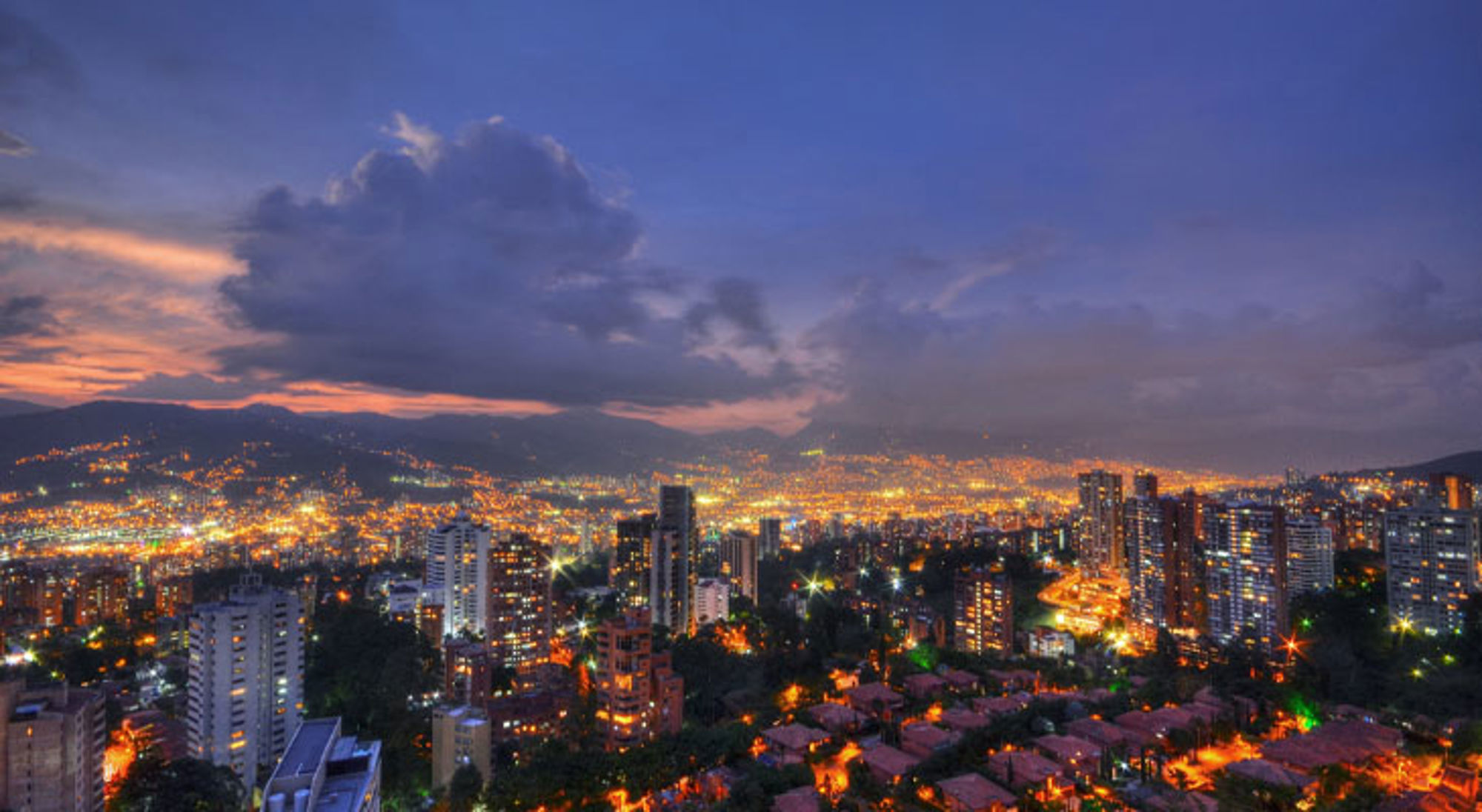 Medellín Avondpanorama van Medellín, Colombia, met verlichte skyline en bergen onder een dramatische wolkenlucht.