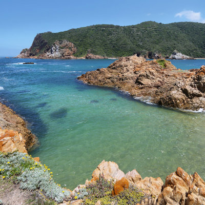 Rotsachtige kust en turquoise baai langs de Tuinroute in Zuid-Afrika, met groene heuvels op de achtergrond.