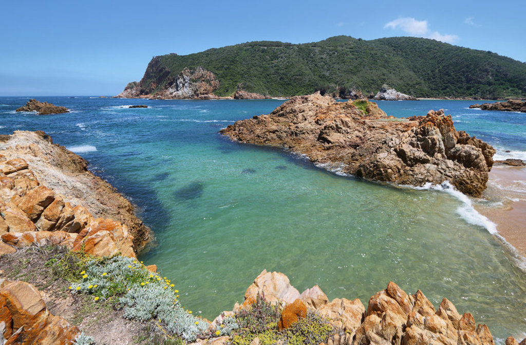 Rotsachtige kust en turquoise baai langs de Tuinroute in Zuid-Afrika, met groene heuvels op de achtergrond.