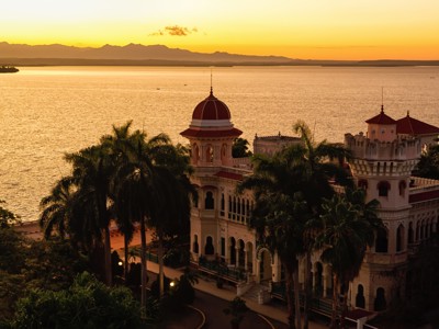Uitzicht op Palacio de Valle in Cienfuegos bij zonsondergang, met palmbomen en de Caribische Zee op de achtergrond.