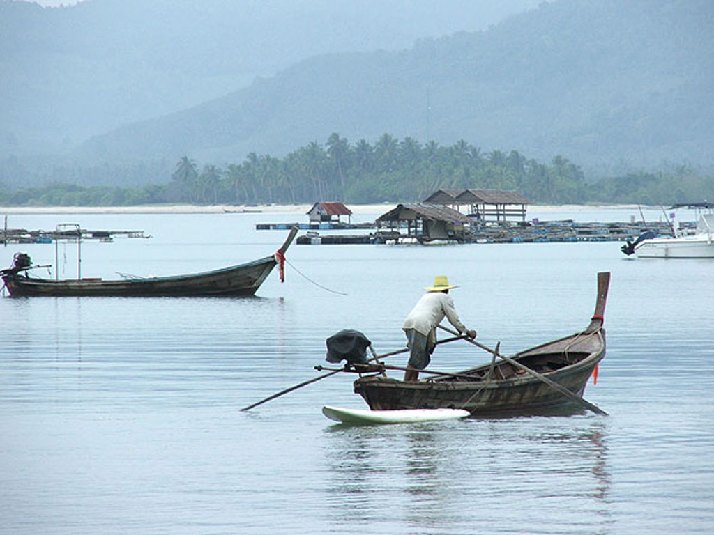 Longtailboot op rustig water in Zuid-Thailand, met vissersdorp en bergen op de achtergrond.