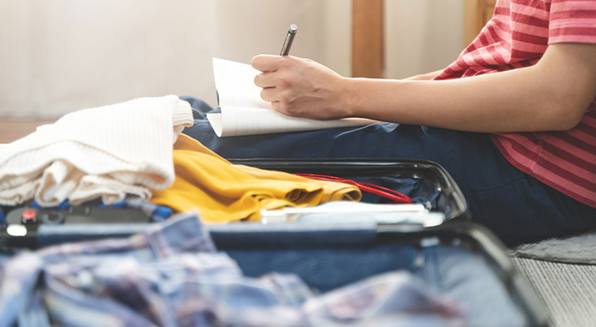 Checklist bagage inpakken Persoon schrijft een paklijst naast een open koffer met kleding, ter voorbereiding op een reis.