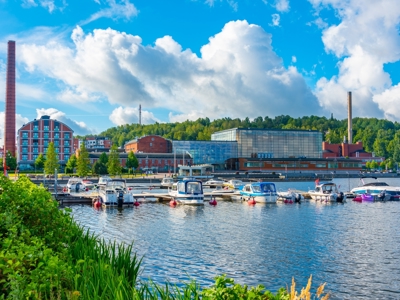 Jachthaven aan de oever van een meer in Scandinavië, met moderne gebouwen en fabrieksschoorstenen op de achtergrond.
