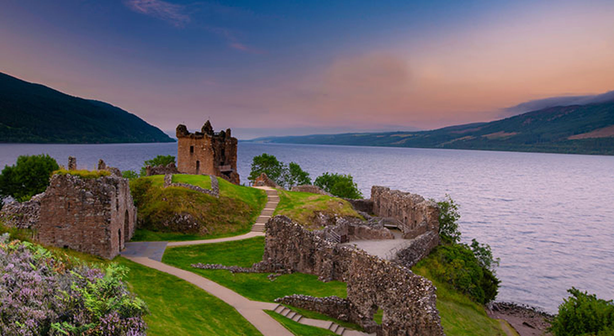 Ruïnes van Urquhart Castle aan Loch Ness bij zonsondergang, met uitzicht over het meer en de heuvels.