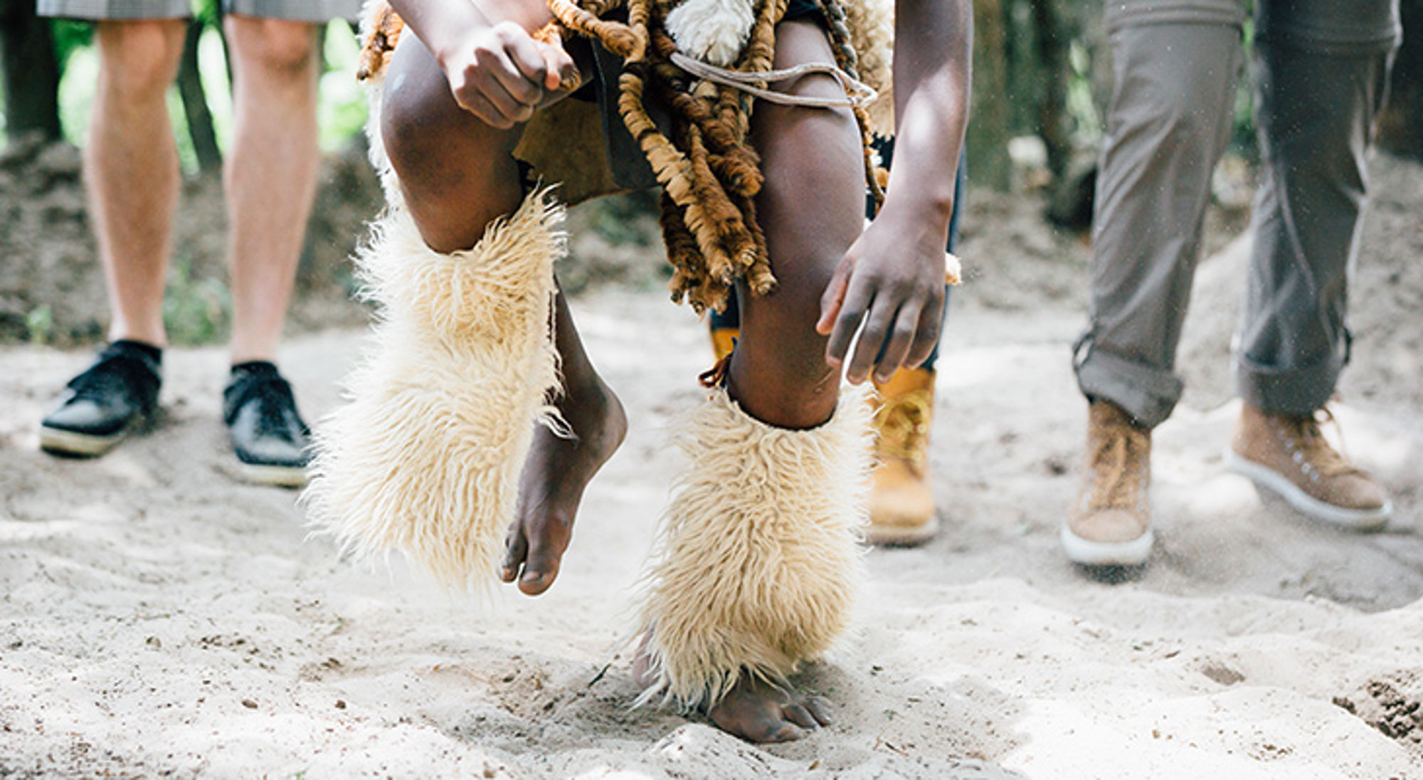 Danser met traditionele beenversiering in zand, met toeschouwers op de achtergrond.