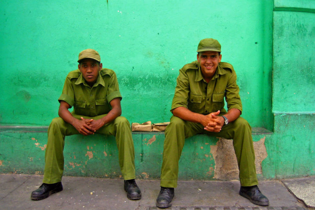 Twee Cubaanse militairen in groen uniform zitten voor een turquoise muur in Havana, Cuba.