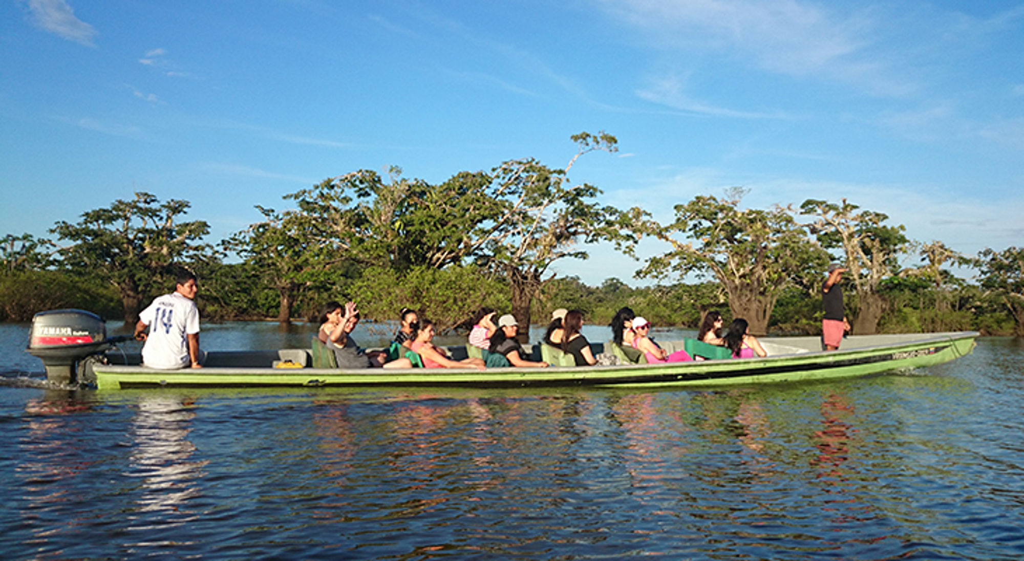 Groep in boot, Cuyabeno, Ecuador Groep reizigers in een lange motorboot op een rivier langs groen mangrovebos onder een blauwe lucht.