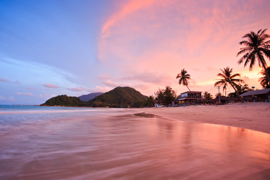 Zonsondergang op een strand in Zuid-Thailand met palmbomen, zee en heuvels op de achtergrond.