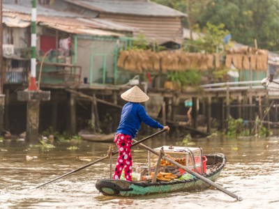 Vrouw met Vietnamese hoed roeit een bootje op de Mekongdelta, langs huizen op palen.