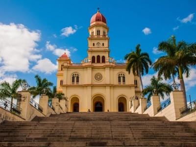 Kerk met klokkentoren en rode koepel in Santiago de Cuba, met palmbomen en brede trap.