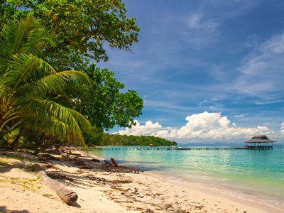 Tropisch strand op Maleisisch Borneo met palmbomen, helderblauw water en een pier naar een paviljoen.