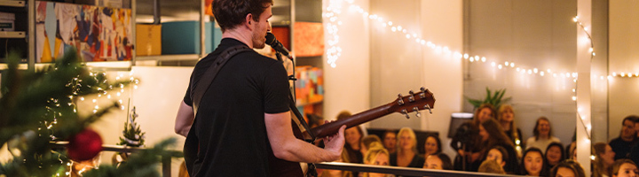 Muzikant speelt gitaar tijdens nieuwjaarsborrel met collega’s, met kerstboom en lichtslingers op de achtergrond.