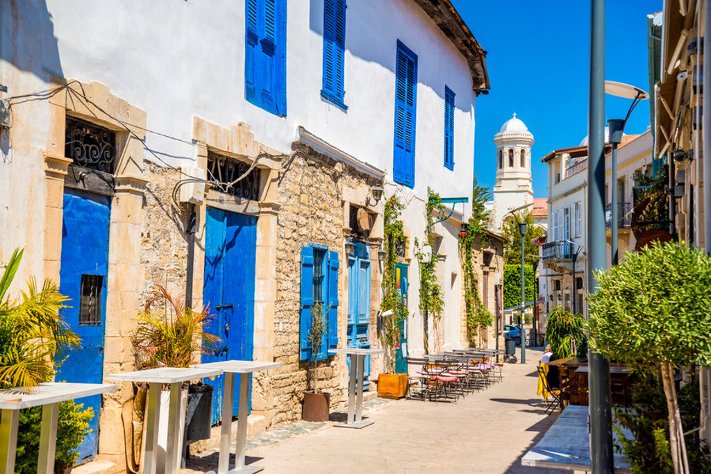 Straatje met witgekalkte huizen en blauwe luiken op Cyprus, met terrasjes en kerktoren in de verte.