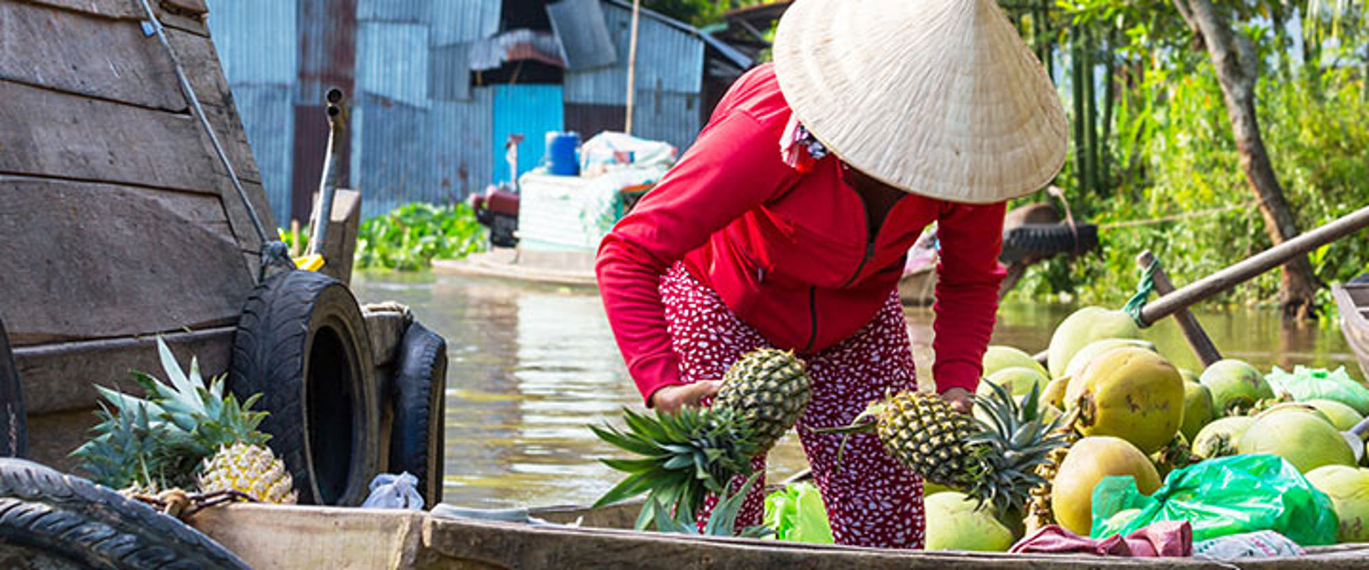 Verkoopster met Vietnamese hoed laadt ananassen en kokosnoten in een boot op een drijvende markt.