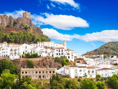 Wit Andalusisch bergdorp met kasteelruïne op rots, omringd door groene heuvels en blauwe lucht.
