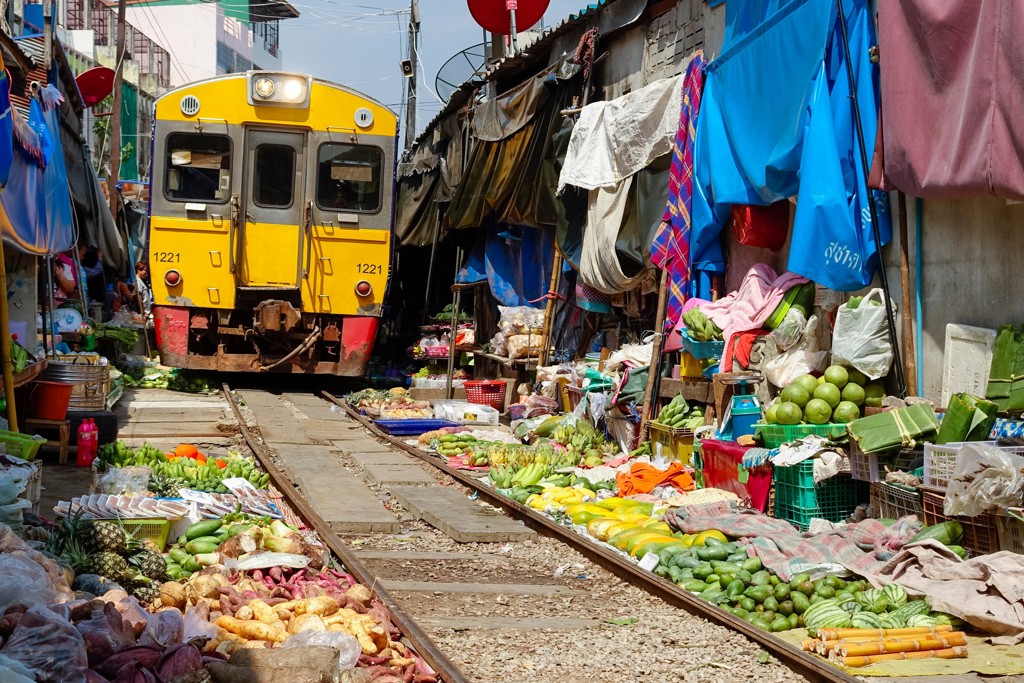 Gele trein langs de Maeklong Railway Market in Thailand, met fruit- en groentekramen direct naast het spoor.