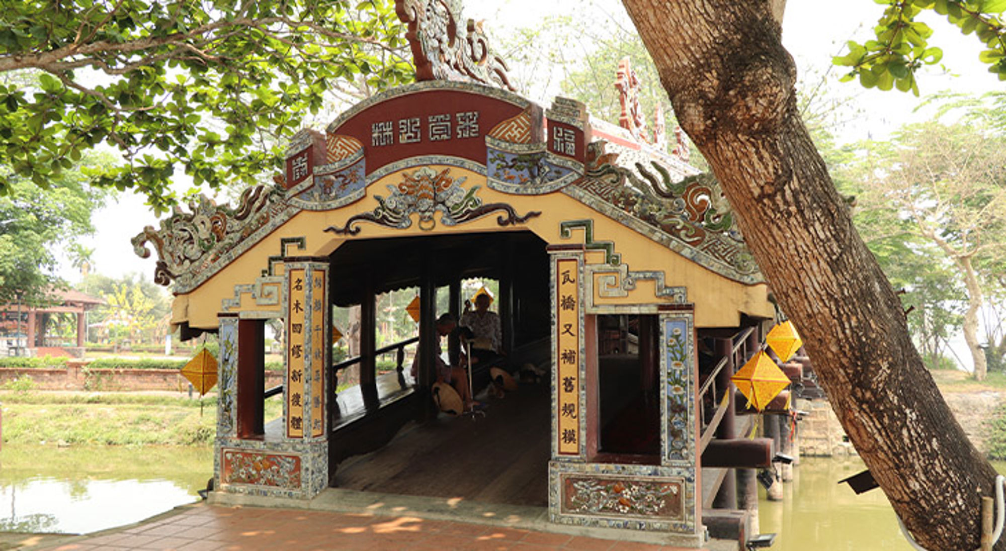 Hué Overdekte, rijk versierde brug met Chinese ornamenten over een kanaal in een park.