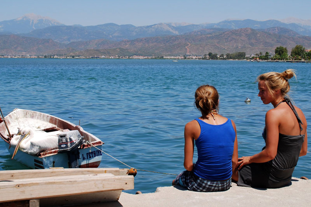Twee reizigers zitten aan de kade bij een bootje met pelikanen, met bergen op de achtergrond in Zuid-Turkije.
