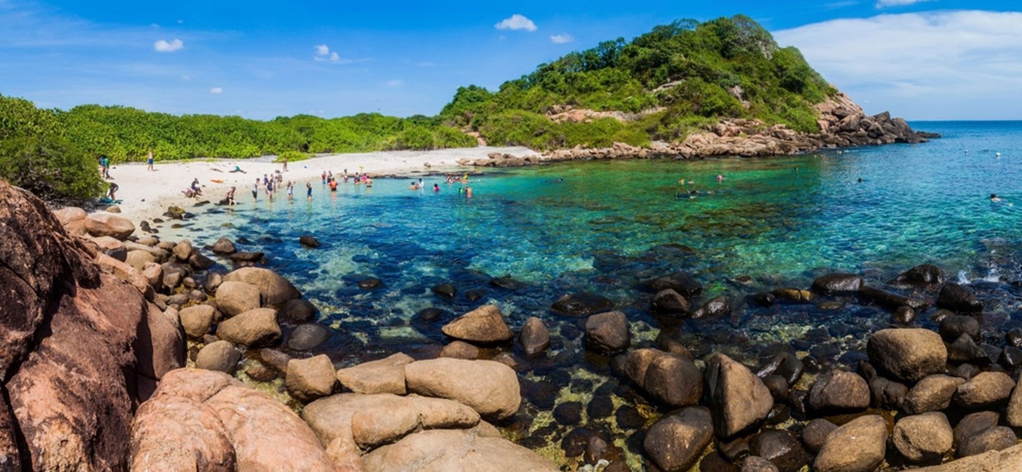 Strandbaai in Sri Lanka met helder turquoise water, rotsen en snorkelaars bij een groene heuvel.