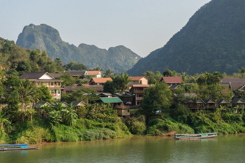Rivierlandschap aan de Mekong in Laos met dorp, longboats en groene karstbergen op de achtergrond.