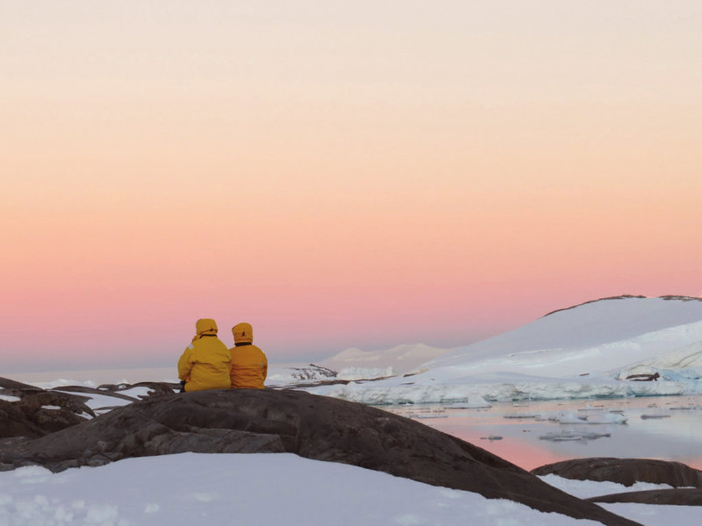 Twee reizigers in gele jassen kijken uit over ijsbergen en besneeuwde kust in Antarctica bij zonsondergang.