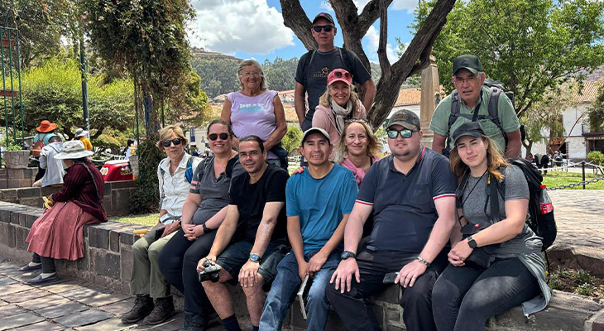 Groepsreis Peru Reisgroep poseert in een park op een stenen muurtje, met bomen en een plein op de achtergrond.