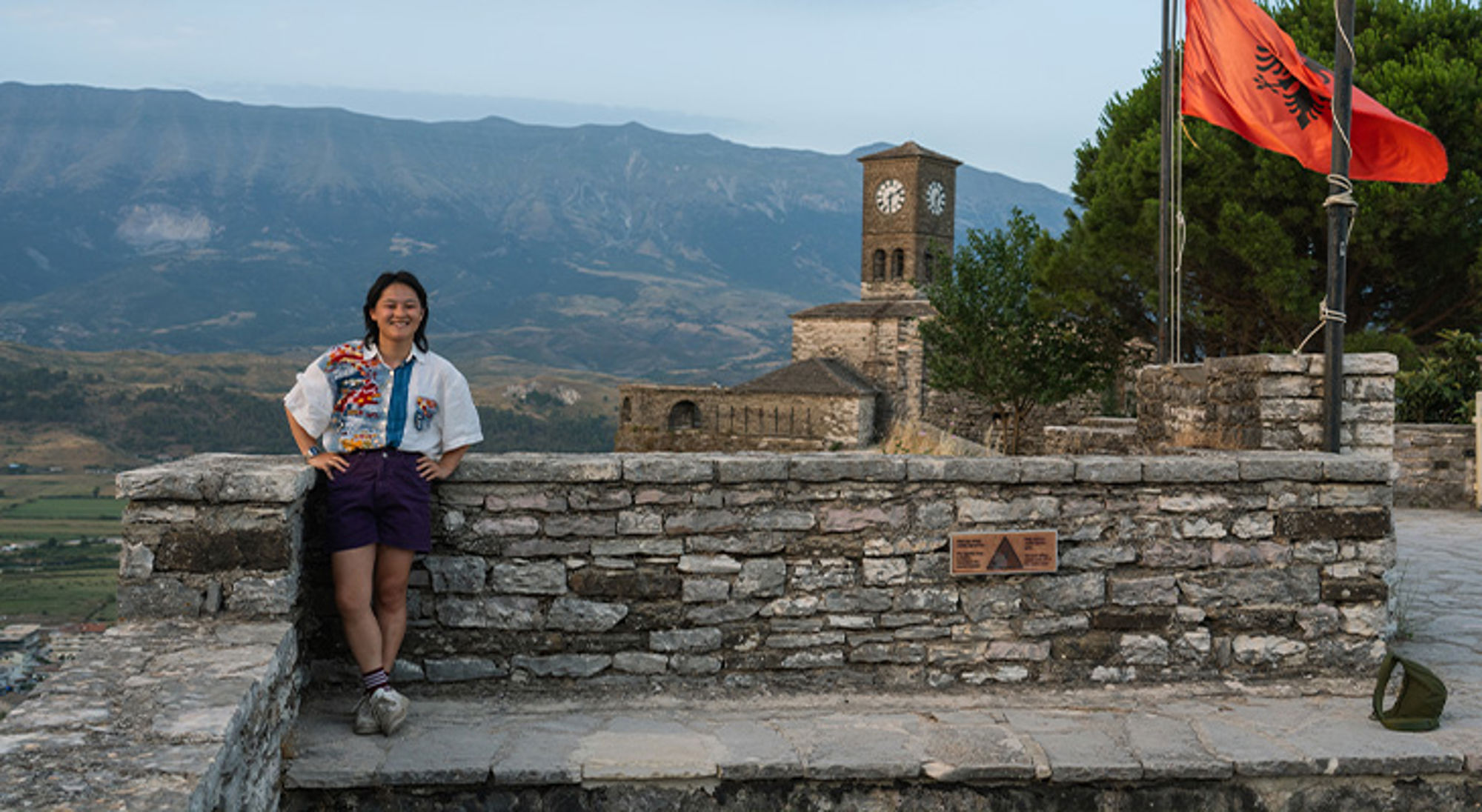Yolan in Albanië Reiziger bij kasteel met Albanese vlag en klokkentoren, met berglandschap op de achtergrond.