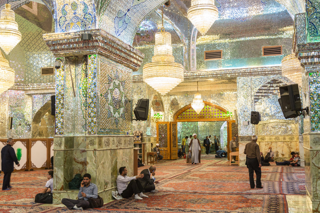 Interieur van een spiegelmozaïekmoskee in Iran met kroonluchters en bezoekers op het tapijt.
