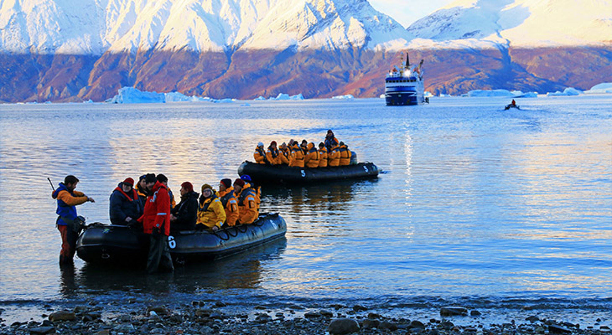 Expeditie Groenland Zodiacboten met passagiers in gele jassen op een ijzige baai, met cruiseschip en besneeuwde bergen op de achtergrond.