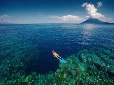 Snorkelaar boven kleurrijk koraal in helderblauw water bij Sulawesi, met vulkaaneiland aan de horizon.