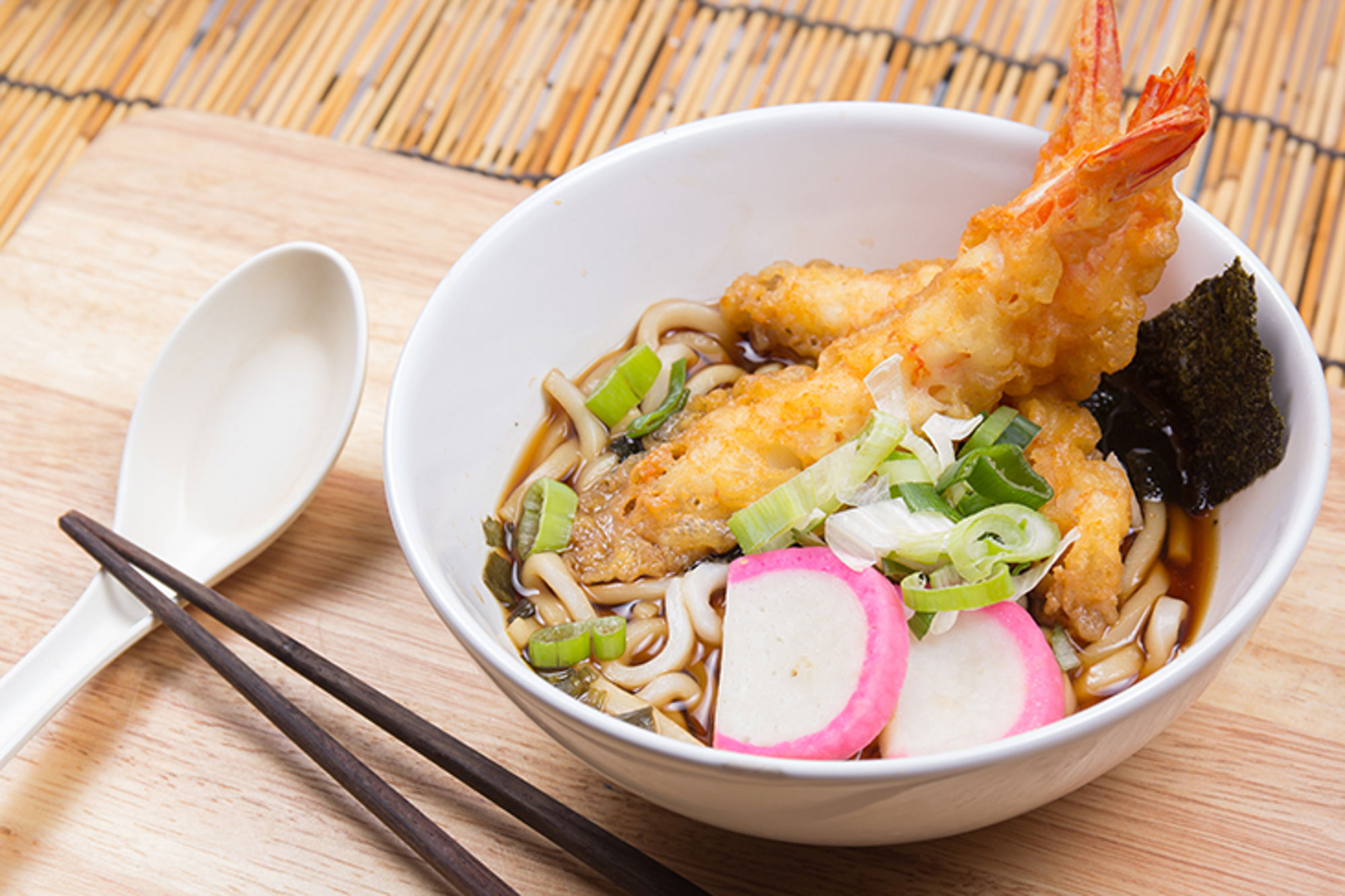 Kom udon noedelsoep met tempura garnaal, lente-ui en nori in een witte kom op houten plank.